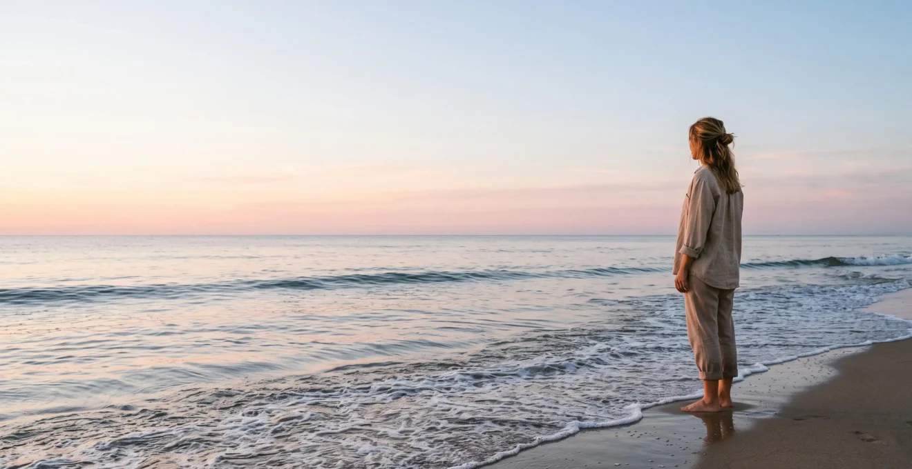 A lone traveler standing at the edge of a calm shoreline at dawn, symbolizing digital disconnection and nervous system reset